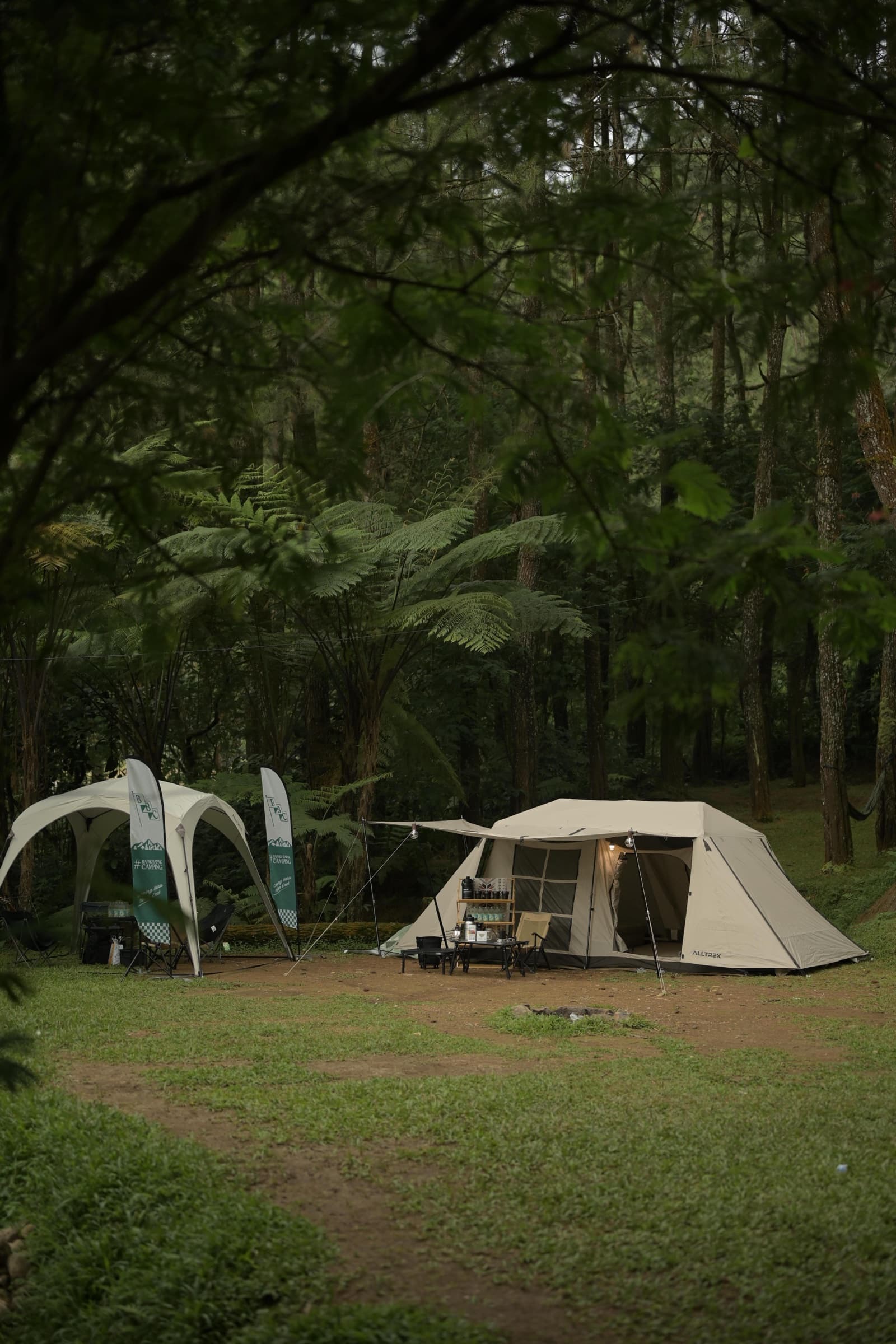 Beige camping tent and outdoor gear display beside a white canopy in a forest clearing